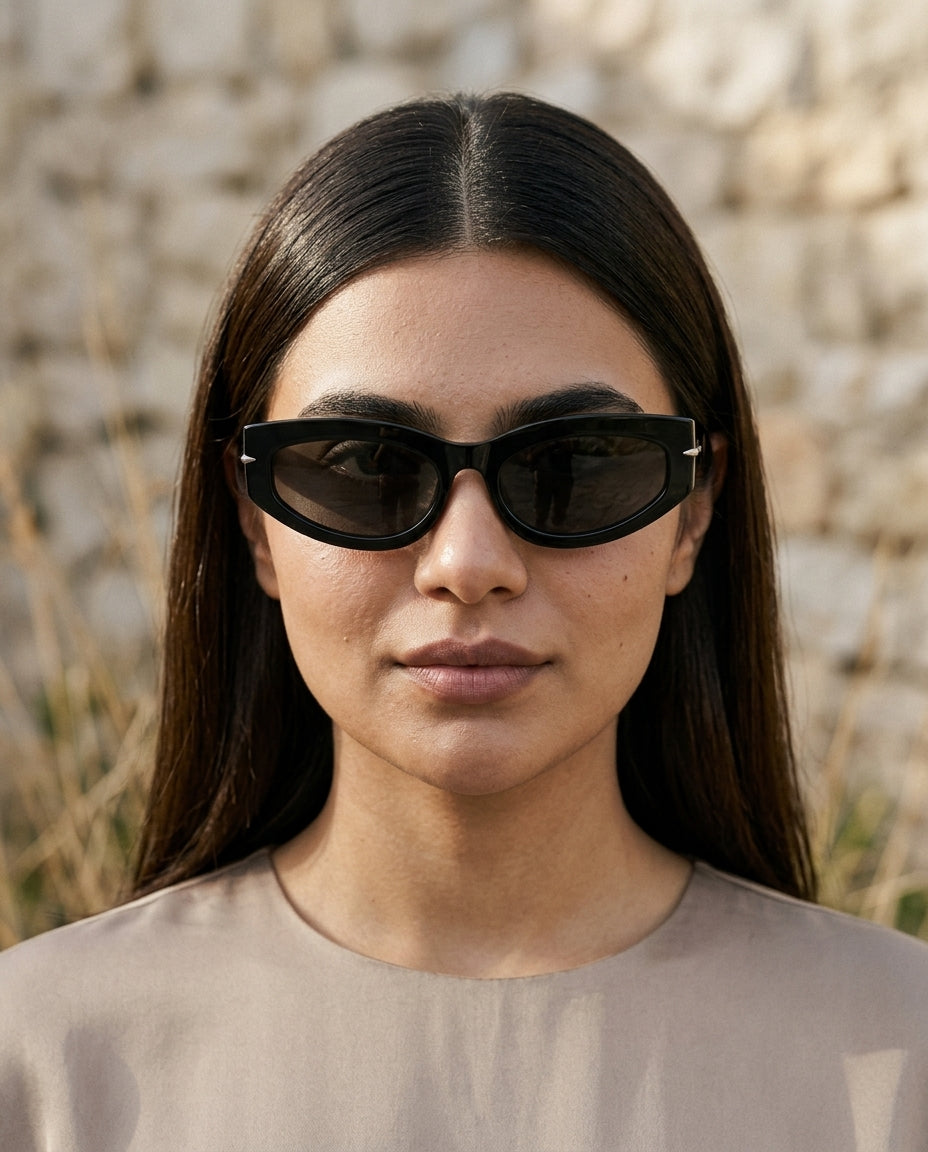 A woman with long dark hair wearing black cat-eye sunglasses and a beige top, photographed against a softly blurred stone wall background.