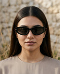 A woman with long dark hair wearing black cat-eye sunglasses and a beige top, photographed against a softly blurred stone wall background.