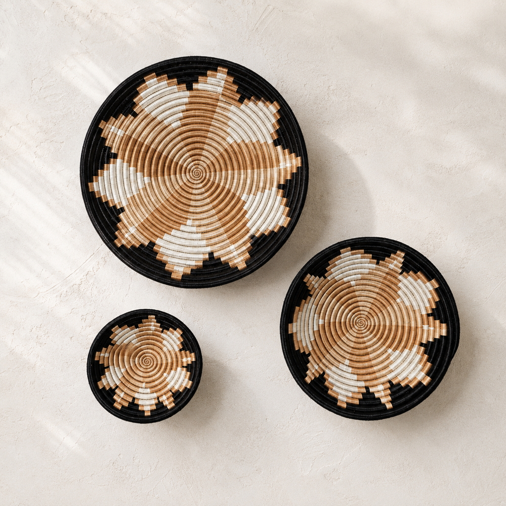 Three round woven baskets in different sizes arranged on a light textured surface. Each basket has a black rim and base with a geometric starburst pattern in alternating black, tan, and off-white sections.