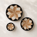 Three round woven baskets in different sizes arranged on a light textured surface. Each basket has a black rim and base with a geometric starburst pattern in alternating black, tan, and off-white sections.