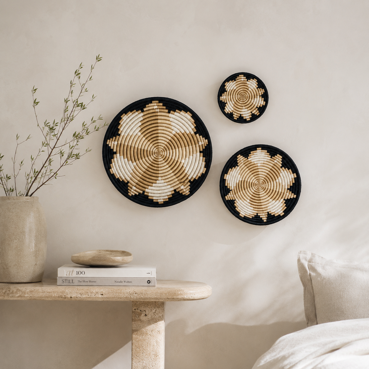 Three round woven wall baskets with spiral patterns in black, beige, and cream, displayed on a light beige wall above a stone side table with books and a vase.