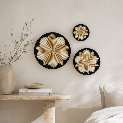 Three round woven wall baskets with spiral patterns in black, beige, and cream, displayed on a light beige wall above a stone side table with books and a vase.