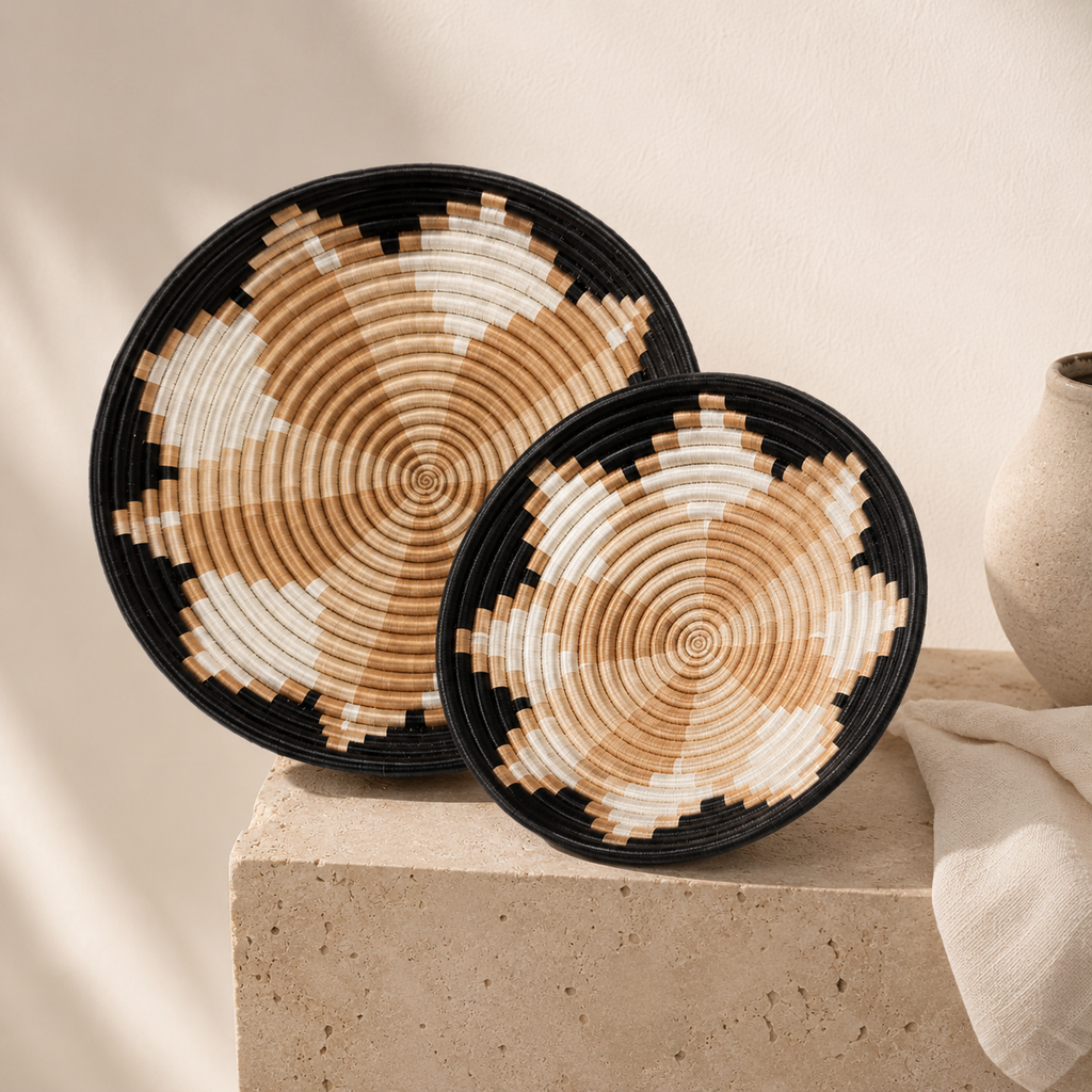 Two round woven baskets with a black rim and a geometric spiral pattern in natural tan and off-white, displayed on a stone pedestal against a warm beige background.