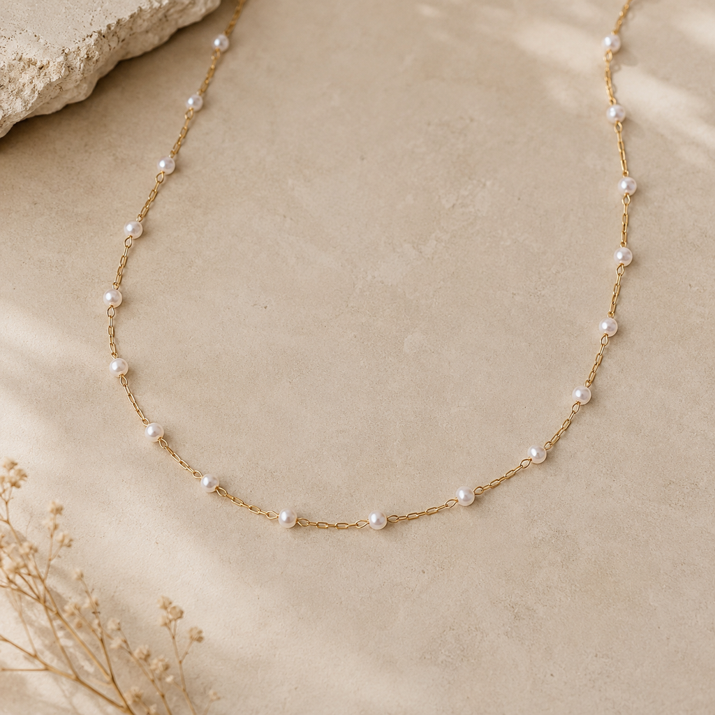 A delicate gold chain necklace adorned with small white freshwater pearls, laid out on a textured beige stone surface with dried flowers in the corner.