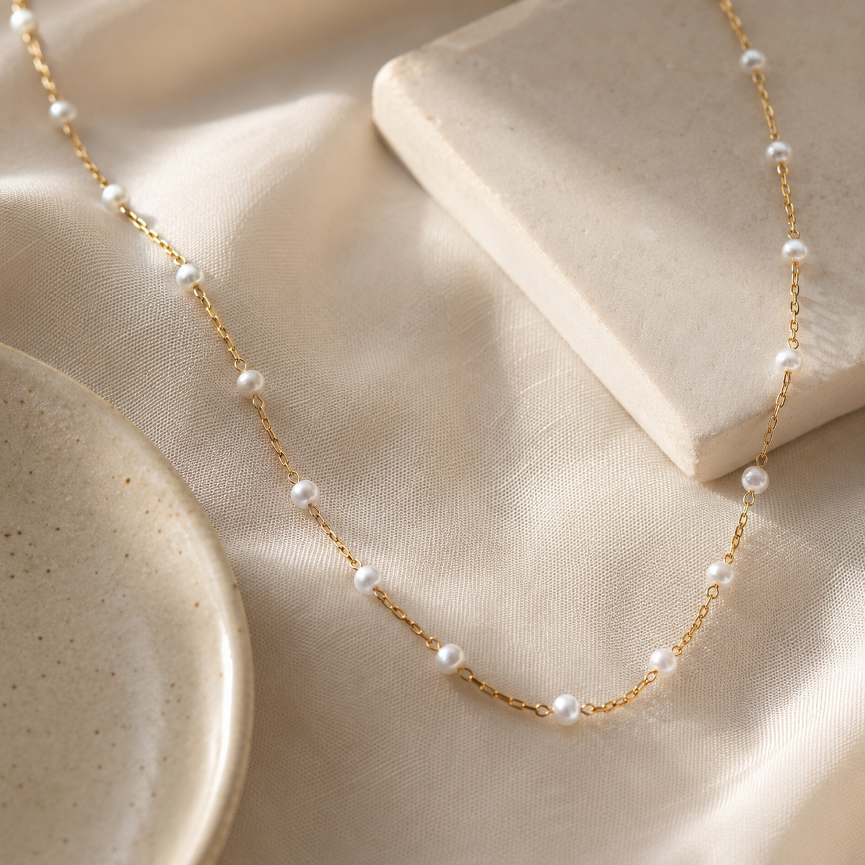 A delicate gold chain necklace adorned with small white pearls, draped elegantly over a cream-colored satin fabric with a stone display block.
