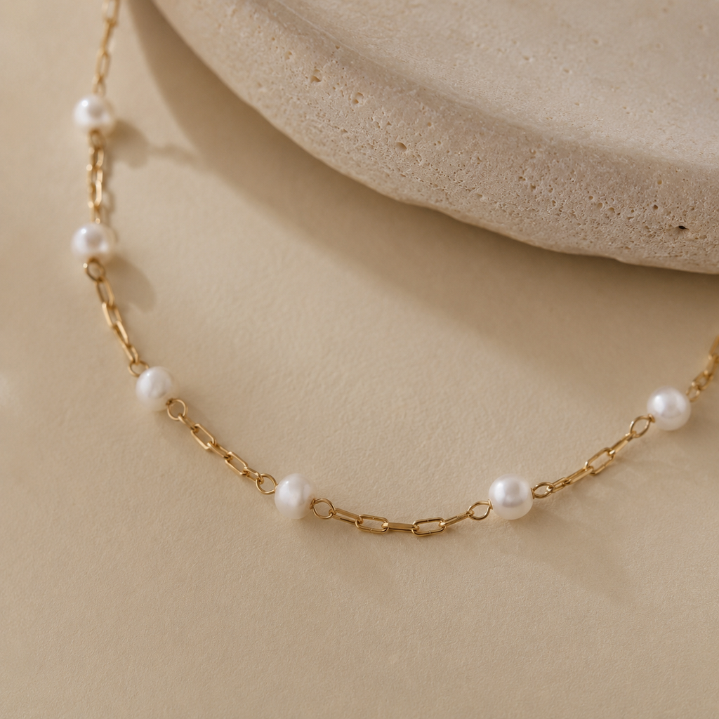 A delicate gold chain necklace with evenly spaced white pearl beads, displayed on a warm beige surface with a textured stone prop in the background.