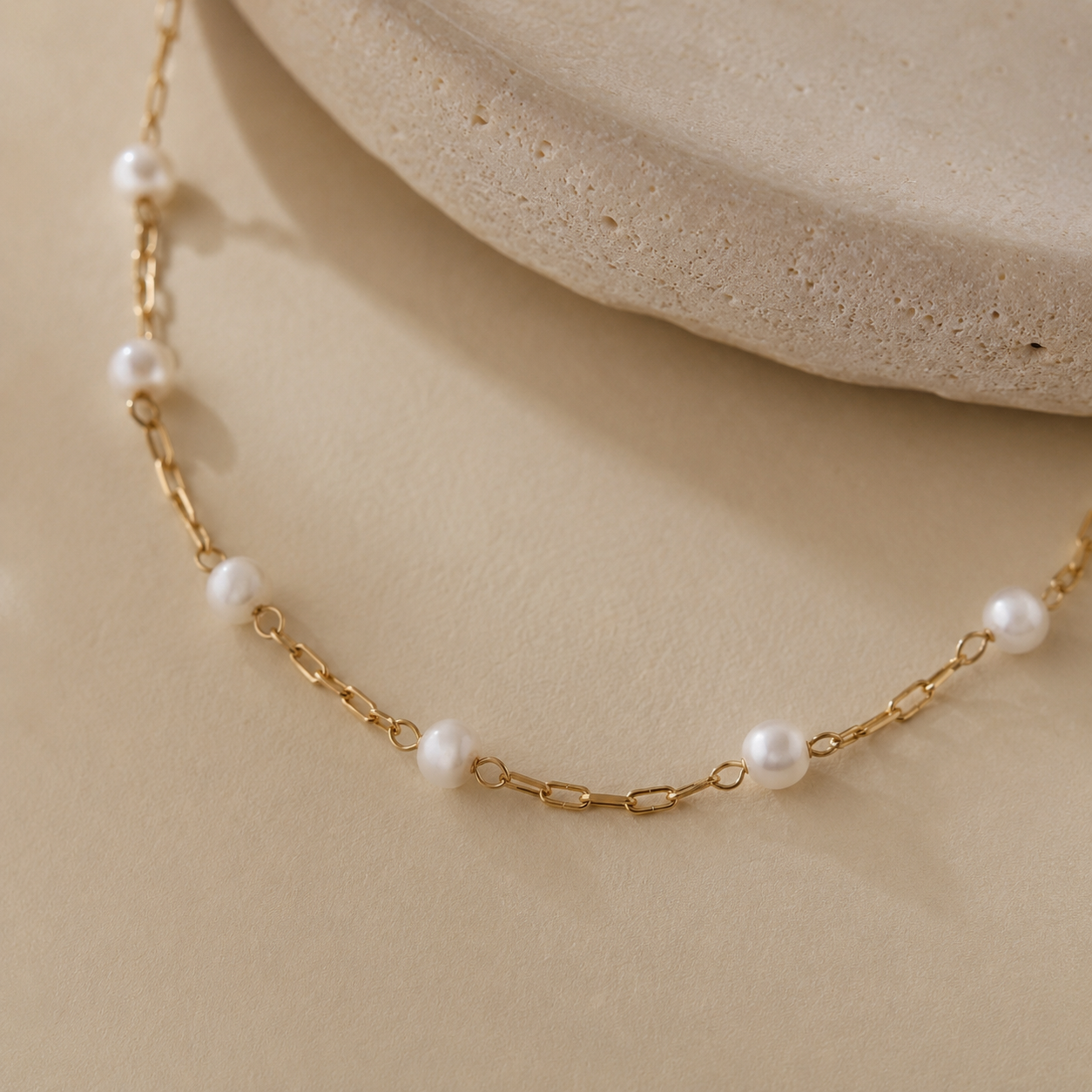 A delicate gold chain necklace with evenly spaced white pearl beads, displayed on a warm beige surface with a textured stone prop in the background.