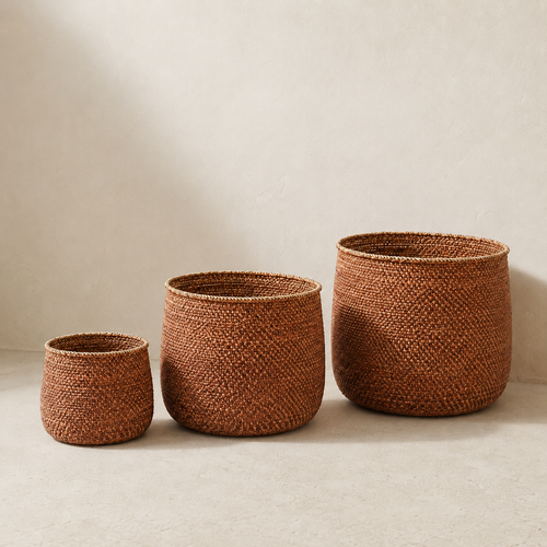 Three woven basket storage bins in graduated sizes, made from natural brown reed or seagrass, arranged on a light beige textured floor against a neutral beige wall.