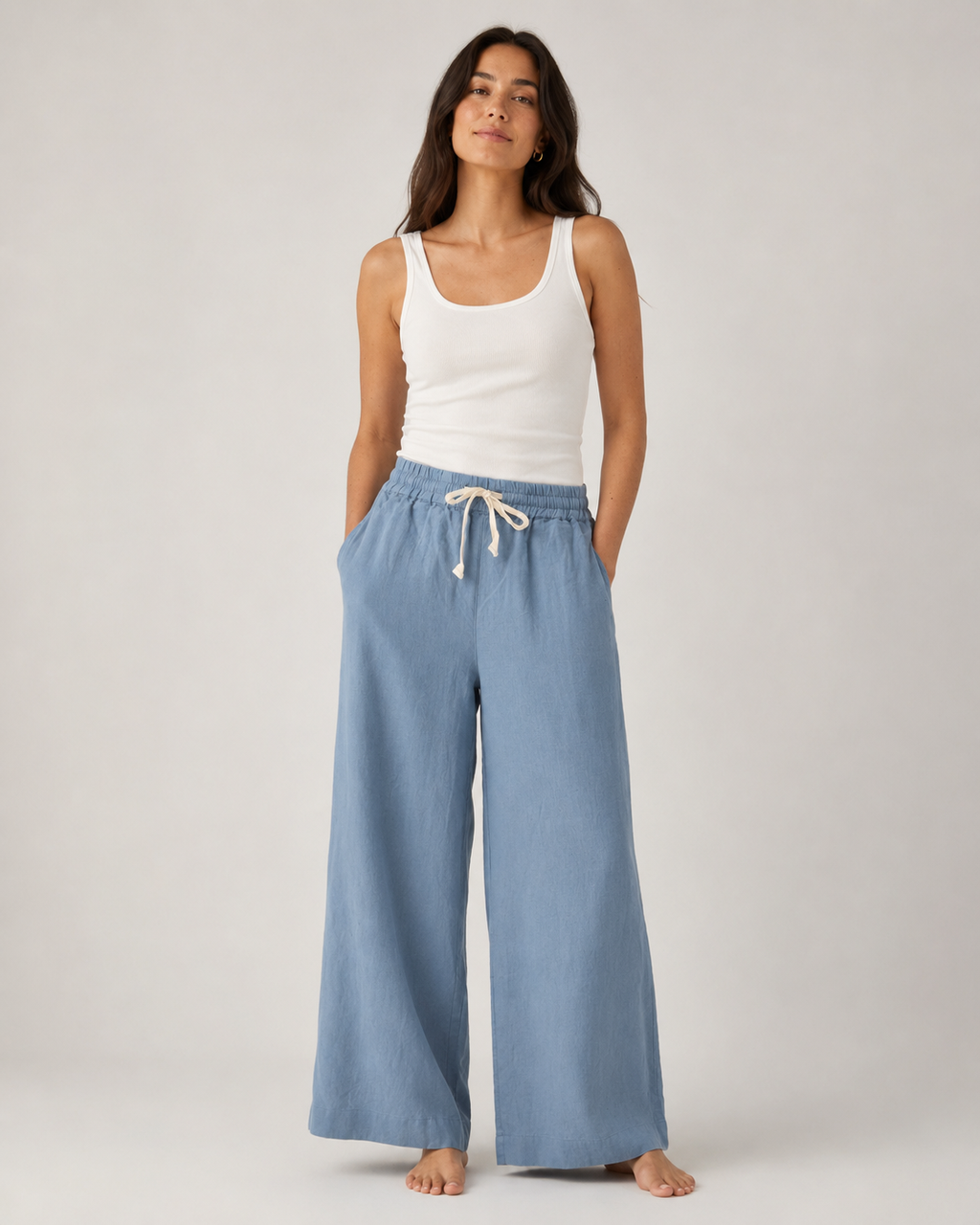 A woman stands barefoot against a light gray studio background, wearing a white ribbed tank top and light blue wide-leg drawstring pants.