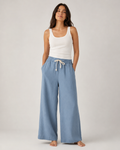 A woman stands barefoot against a light gray studio background, wearing a white ribbed tank top and light blue wide-leg drawstring pants.