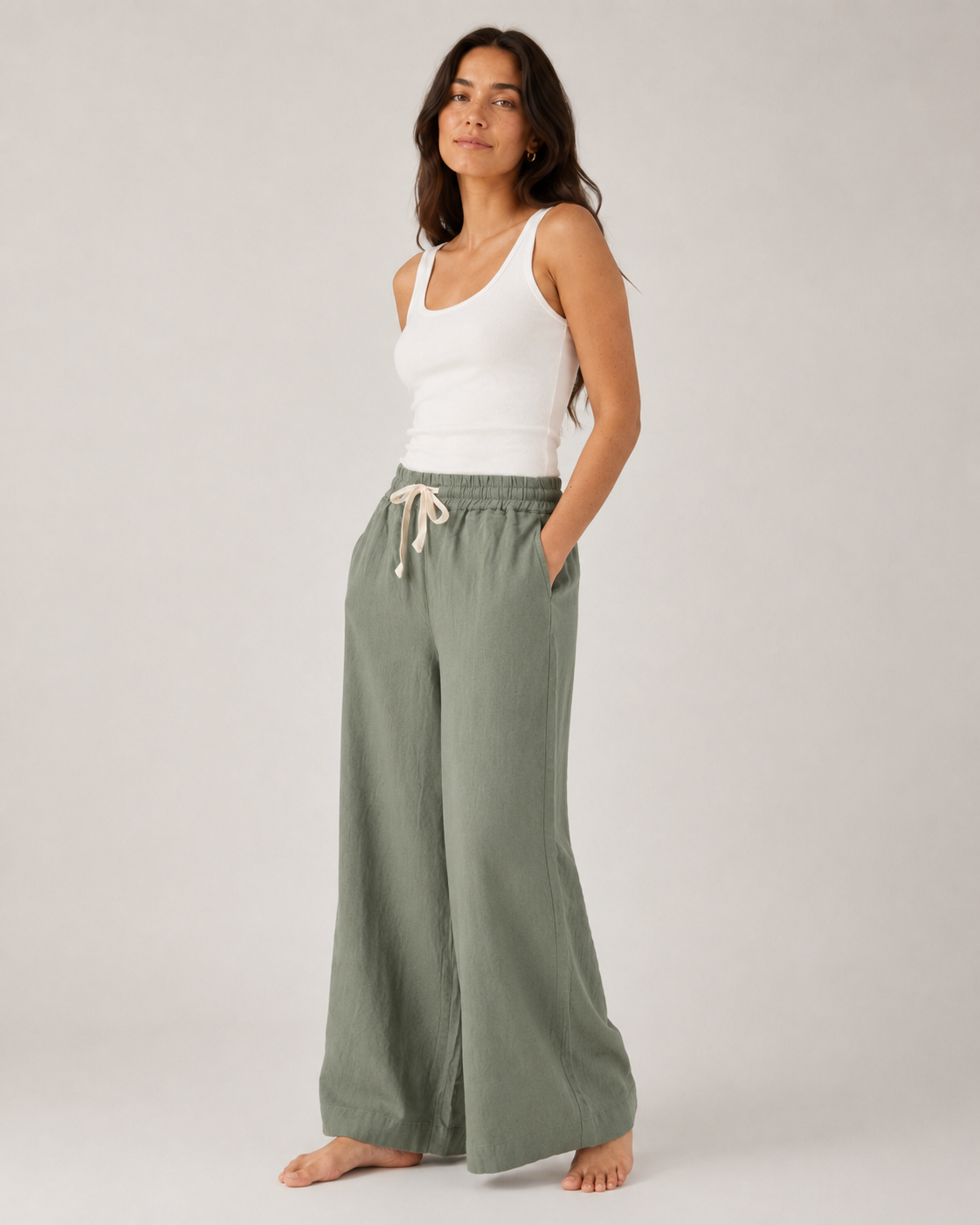 A woman modeling wide-leg sage green lounge pants with a white drawstring waist, paired with a white tank top, against a plain light gray studio background.