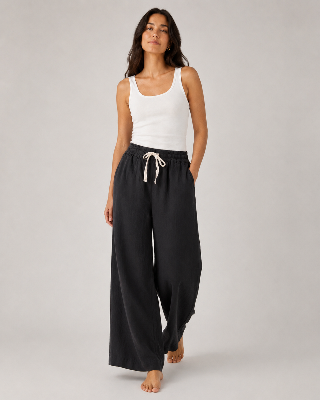 A woman modeling black wide-leg lounge pants with a white drawstring, paired with a white tank top, against a light gray studio background.