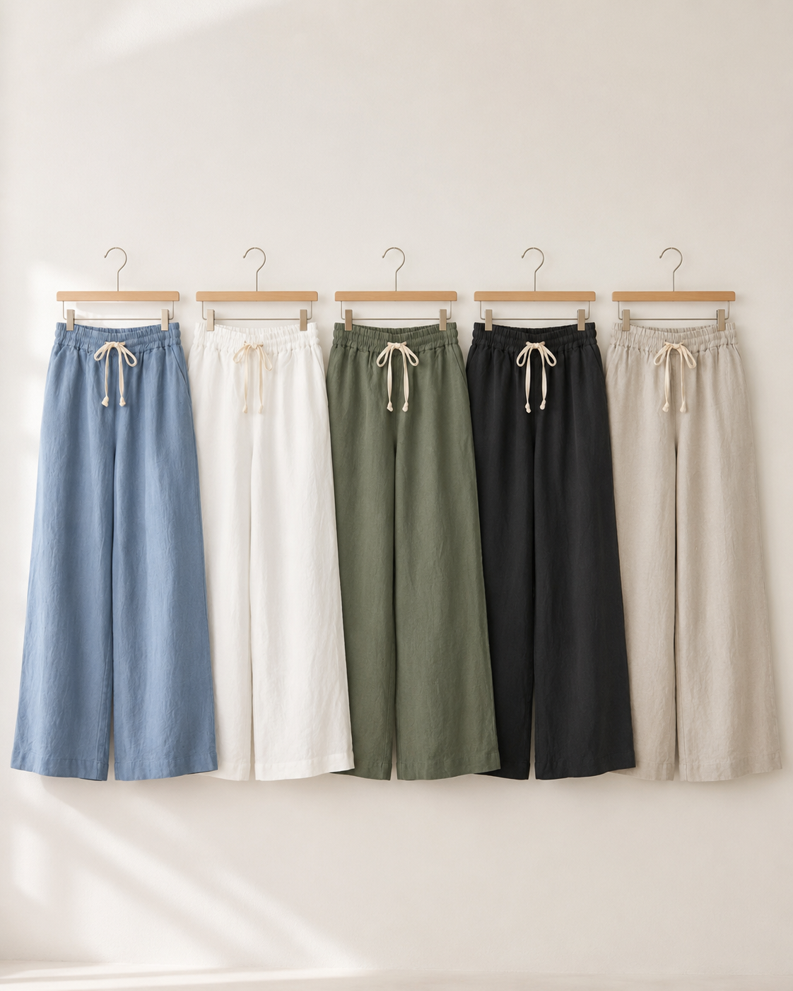 Five pairs of wide-leg drawstring pants in light blue, white, sage green, black, and beige, hanging on wooden hangers against a warm off-white wall.