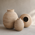 Three woven natural-fiber decorative vases in different shapes and sizes, arranged on a light concrete surface against a neutral textured wall.