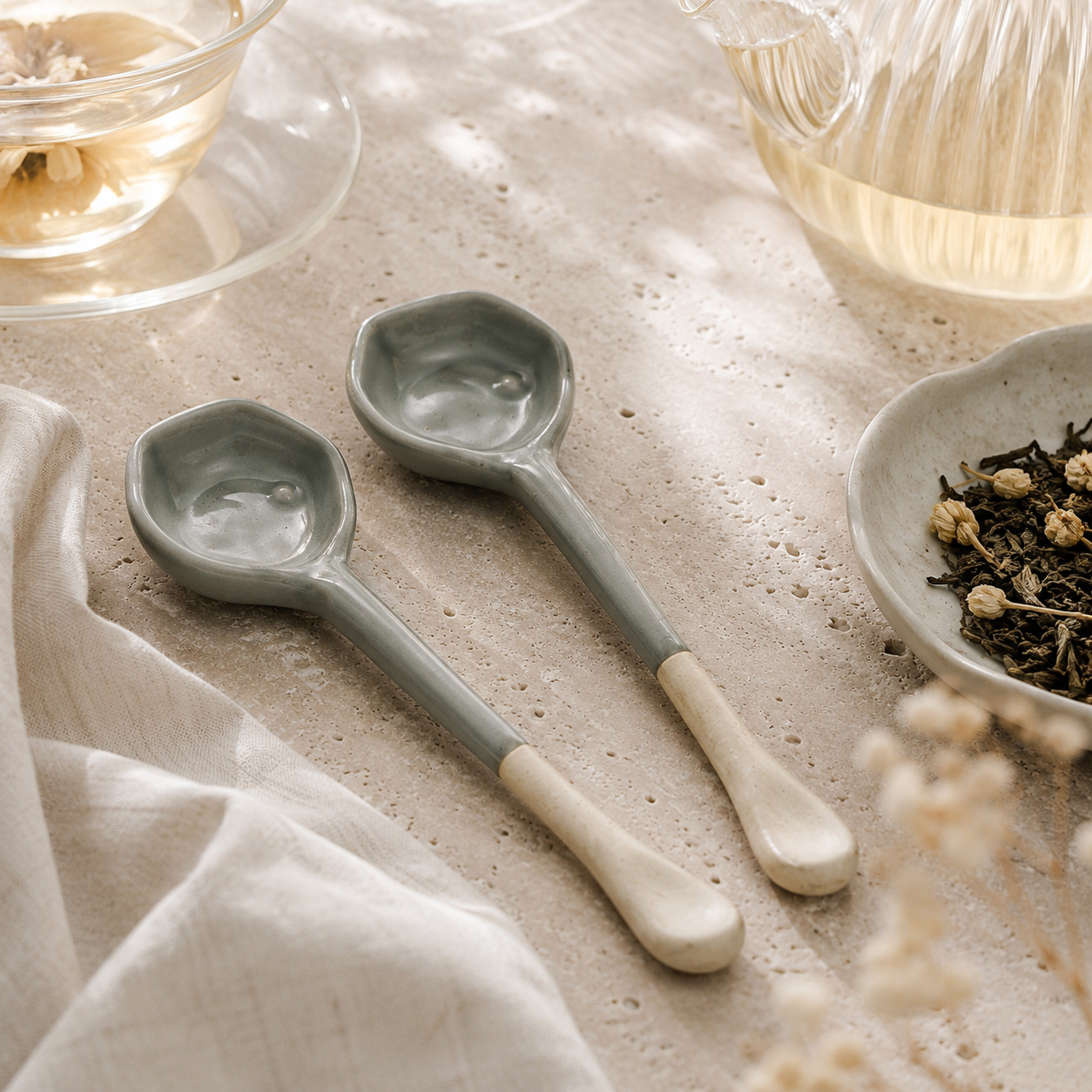 Two ceramic tea spoons with muted sage green bowls and cream handles, styled on a textured beige surface with tea and dried flowers.