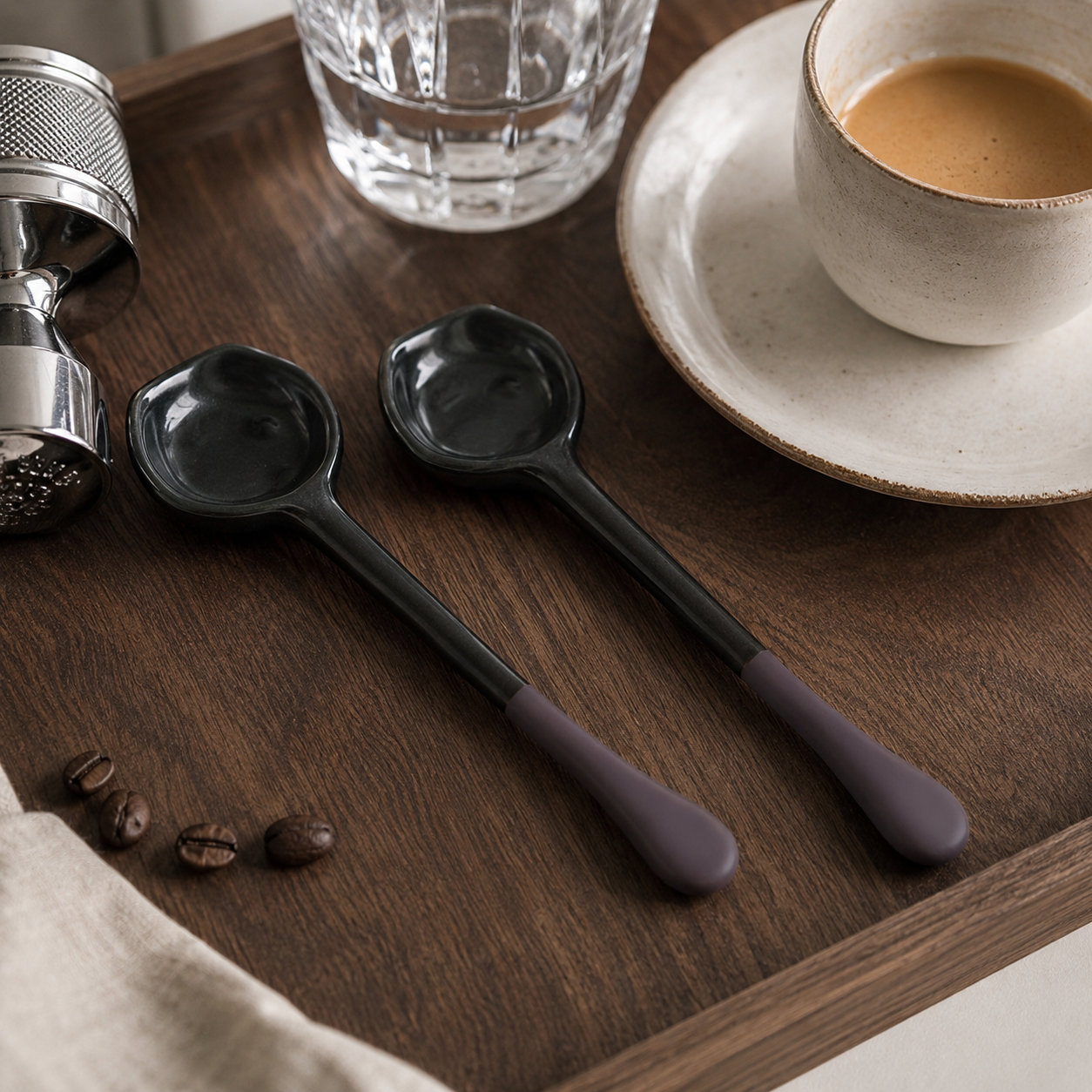Two ceramic spoons with matte black heads and muted purple handles resting on a dark wooden tray, styled with coffee beans, a glass of water, a metal grinder, and a cup of coffee on a saucer.