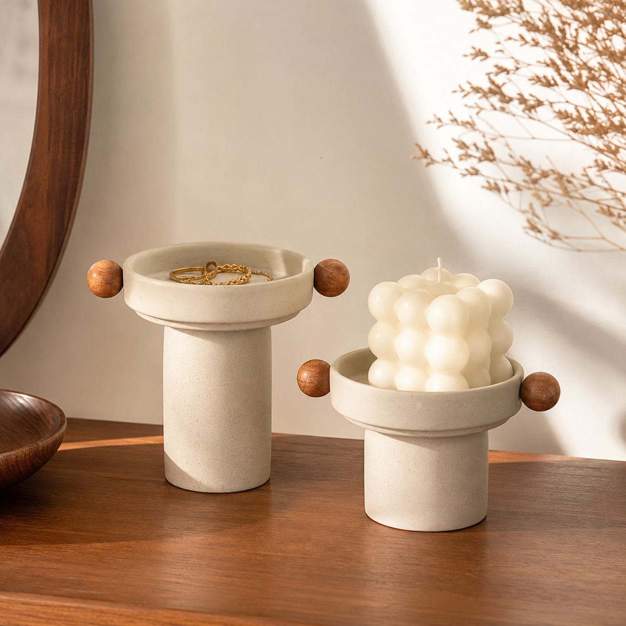 Two cream-colored ceramic candle holders with wooden ball accents sit on a wooden table. One holder is empty, and the other holds a white bubble-style candle.