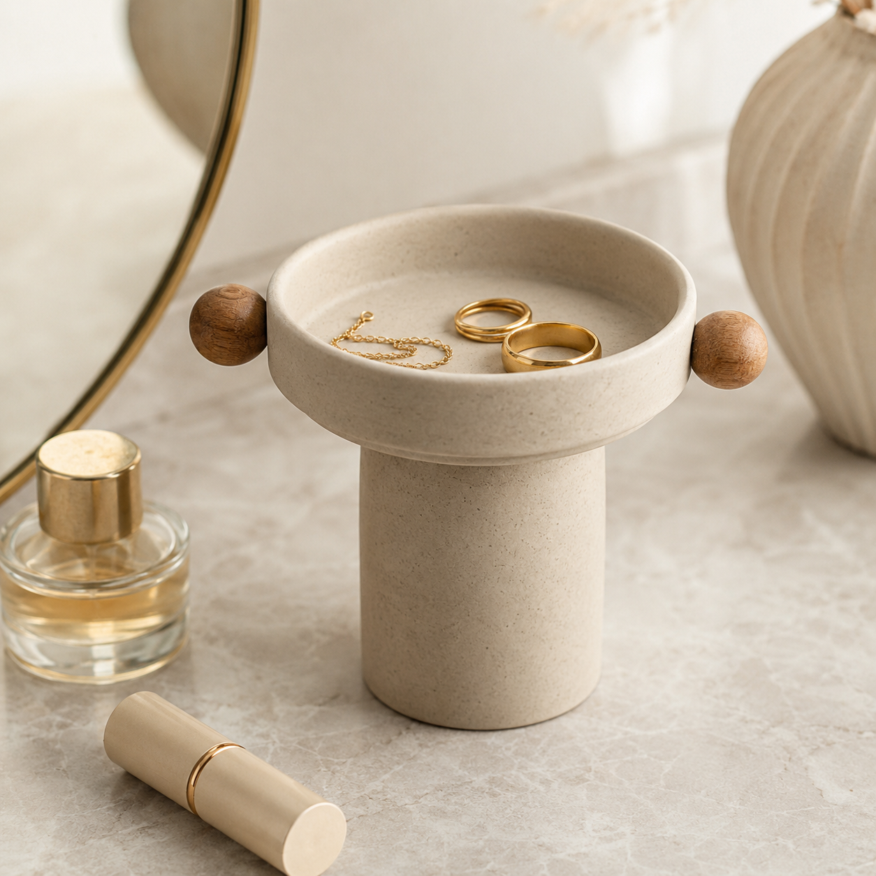A small cylindrical jewelry organizer tray with a round top, wooden side knobs, and gold rings and a chain displayed inside, styled on a light stone surface with a perfume bottle, lipstick, and a vase in the background.
