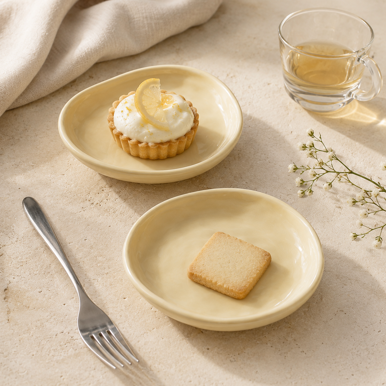 Two round cream-colored ceramic plates styled on a beige textured surface, with a small tart on the upper plate and a square cookie on the lower plate, alongside a fork, a glass of tea, and delicate flowers.