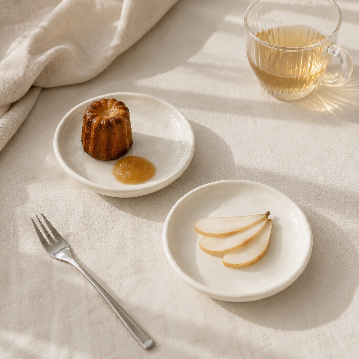 Two small round white ceramic plates styled on a beige linen tablecloth with a silver fork, a small cake, a dollop of jam, and slices of pear. A glass of tea sits in the background.
