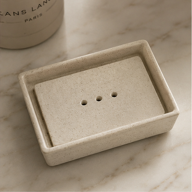A four-panel collage showing a rectangular soap dish with a matching lid in a warm neutral stone finish. The soap dish is shown empty and with a bar of soap resting inside, styled on a marble surface, a wooden tray with a brush, and next to a folded towel.
