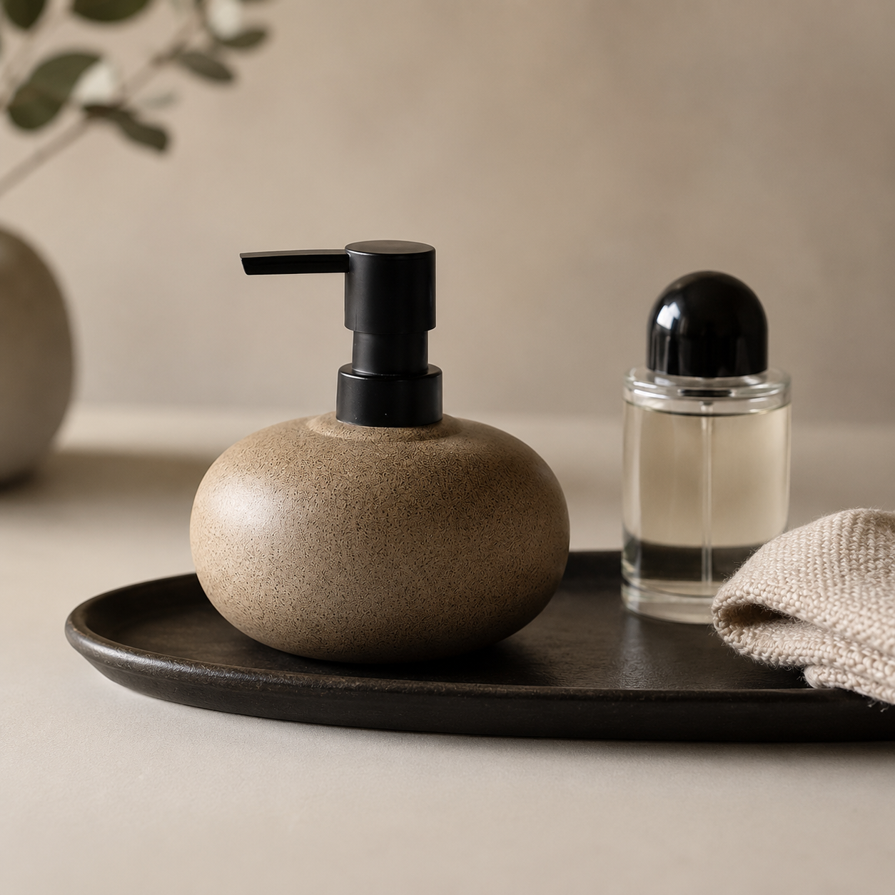 A round beige soap dispenser with a matte black pump sits on a dark tray next to a small clear bottle and a folded beige cloth, against a soft neutral background.