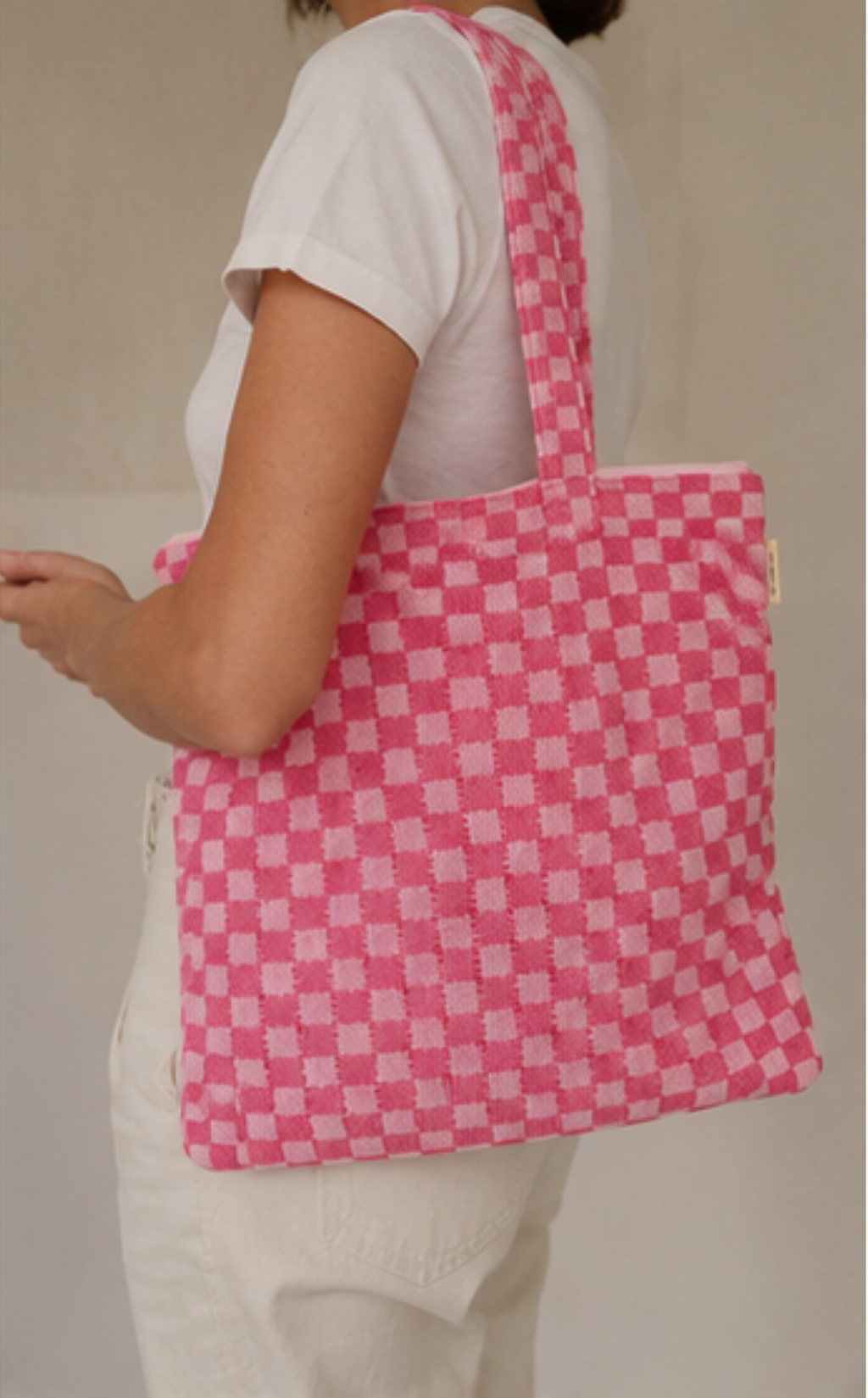 A pink and white checkered canvas tote bag with a single shoulder strap, worn by a person wearing a white t-shirt and white pants against a neutral beige background.