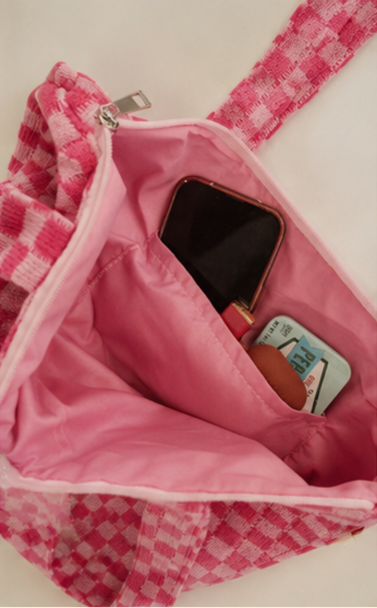 A close-up of a pink and white checkered bag shown open, revealing a pink interior with a black smartphone and a small card or wallet inside.