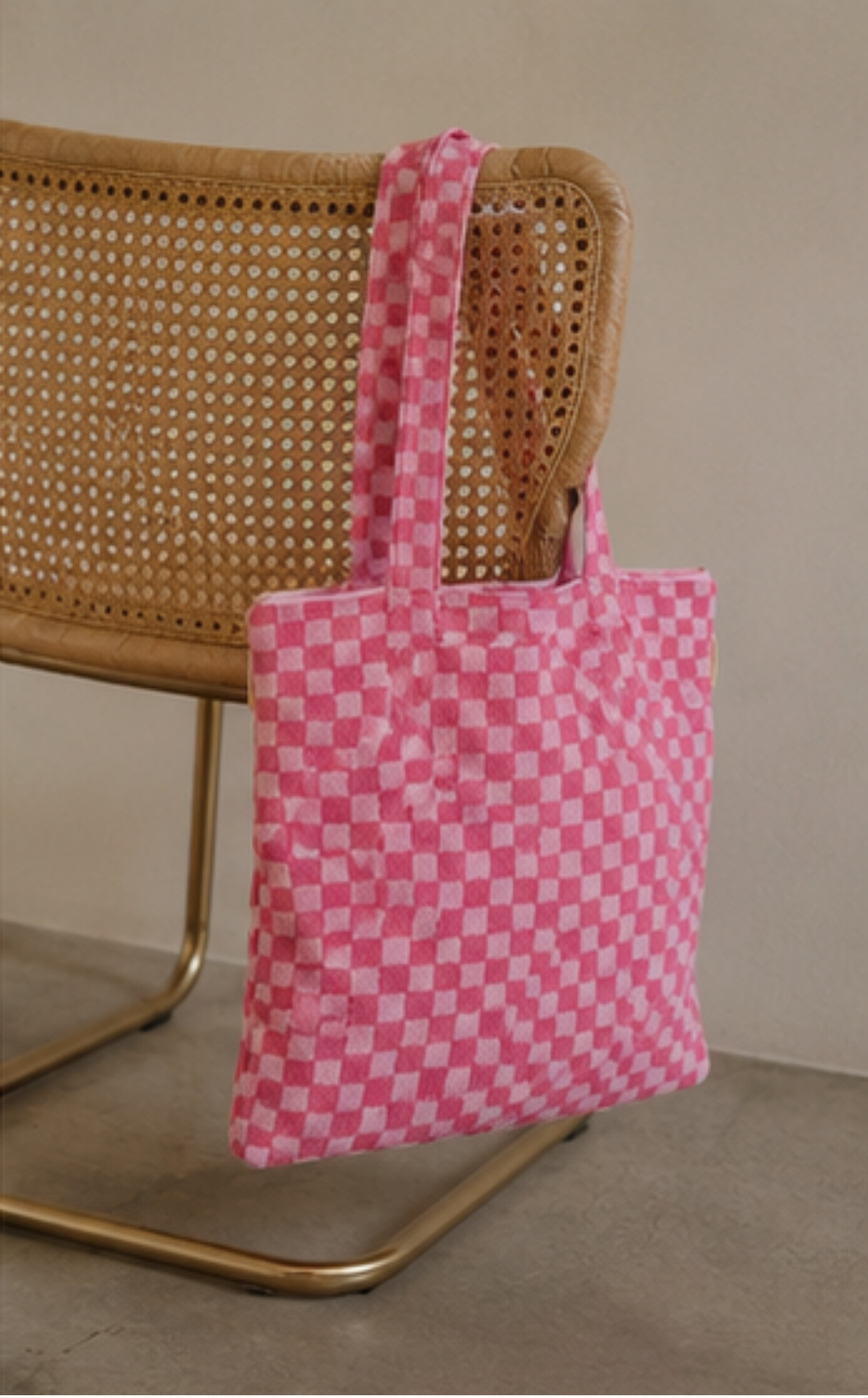 A pink and white checkered canvas tote bag hanging from a woven cane chair with a gold metal frame, set against a neutral beige wall and concrete floor.