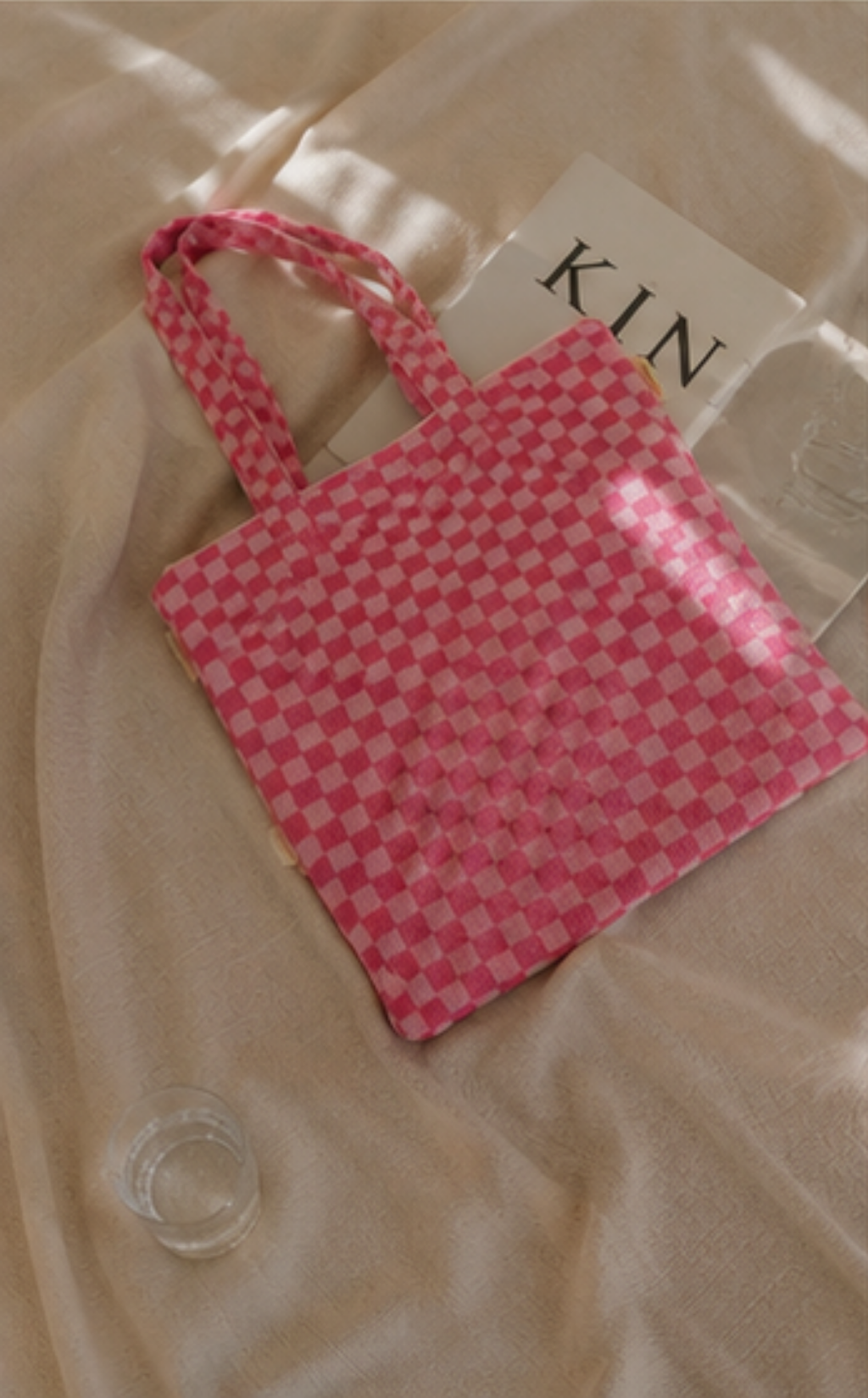 A pink and white checkered fabric tote bag with two handles, resting on a beige satin-like fabric surface next to a white card with the letters 'KIN'.