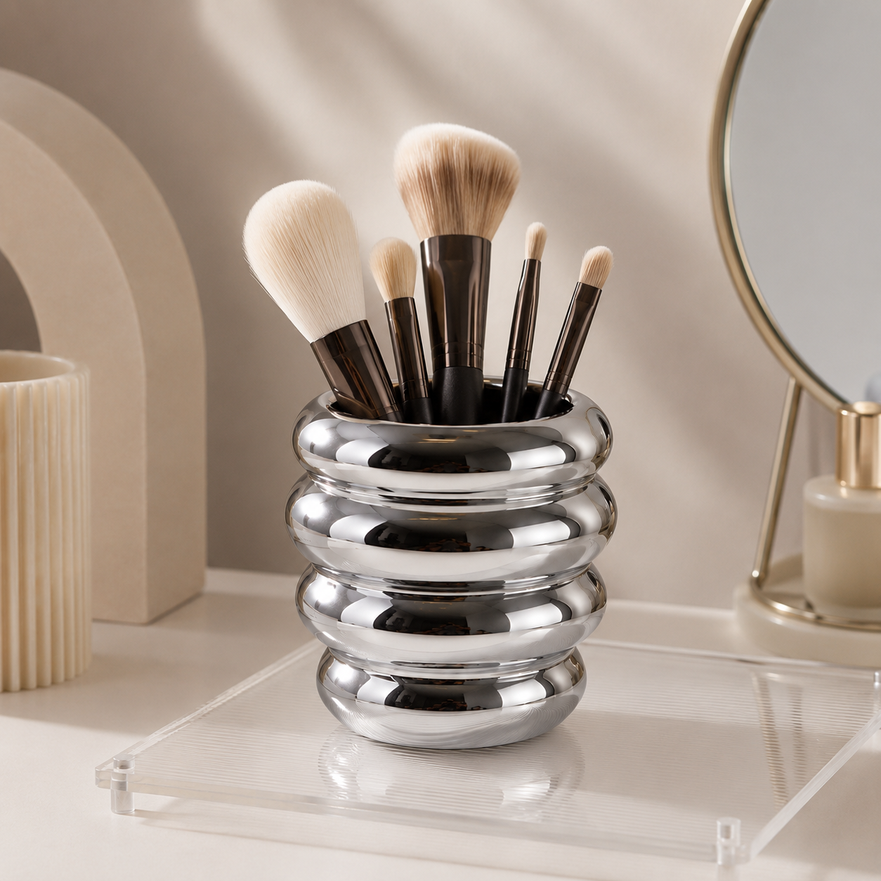 A polished silver metallic makeup brush holder filled with makeup brushes, styled on a clear acrylic tray on a light beige surface with soft, warm lighting.