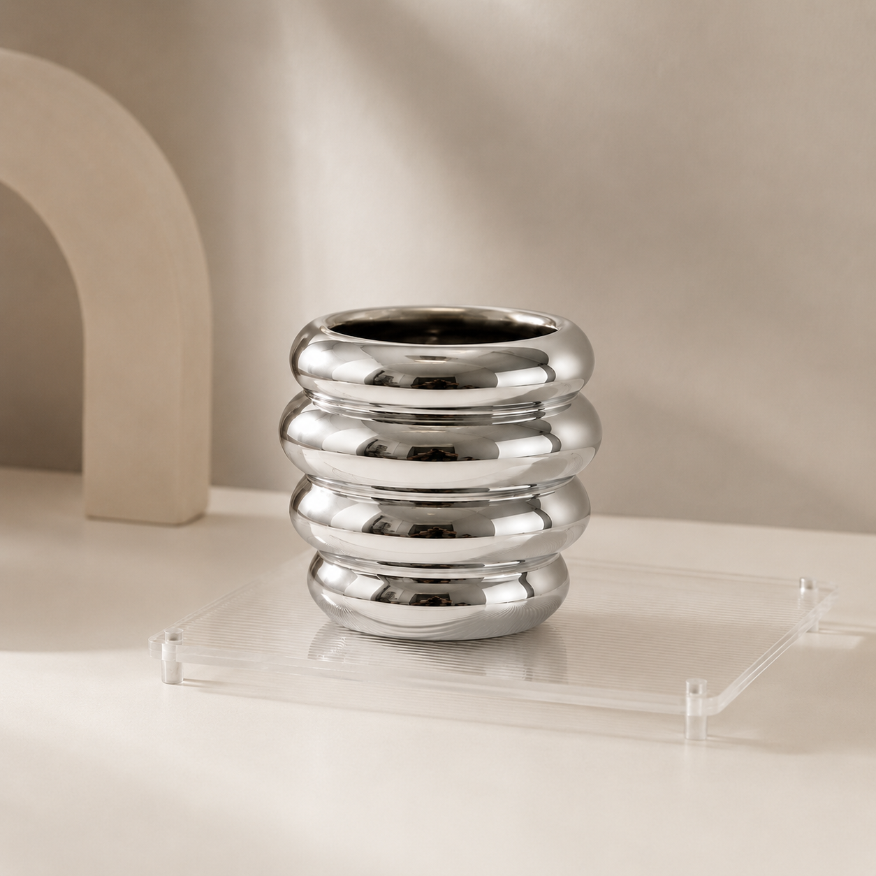 A polished silver metallic vase with a stacked, ribbed design, displayed on a clear acrylic riser against a warm beige background.