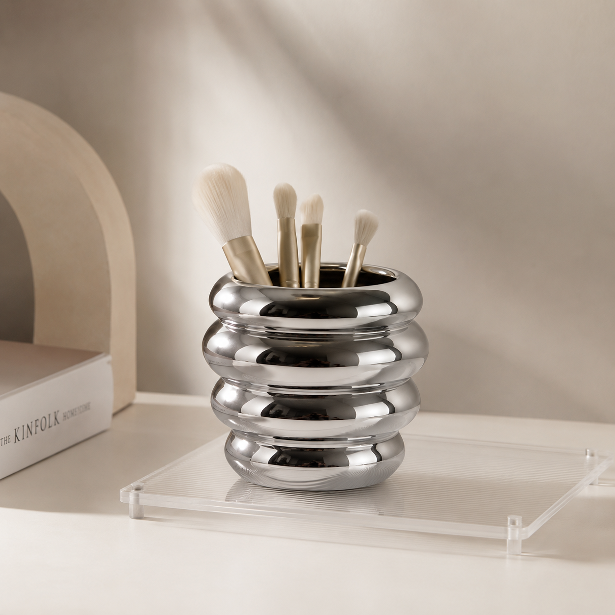 A shiny silver metallic makeup brush holder with a ribbed, rounded design, holding four makeup brushes. The holder is placed on a clear acrylic tray on a light beige surface, with a neutral beige background and a decorative book on the left.