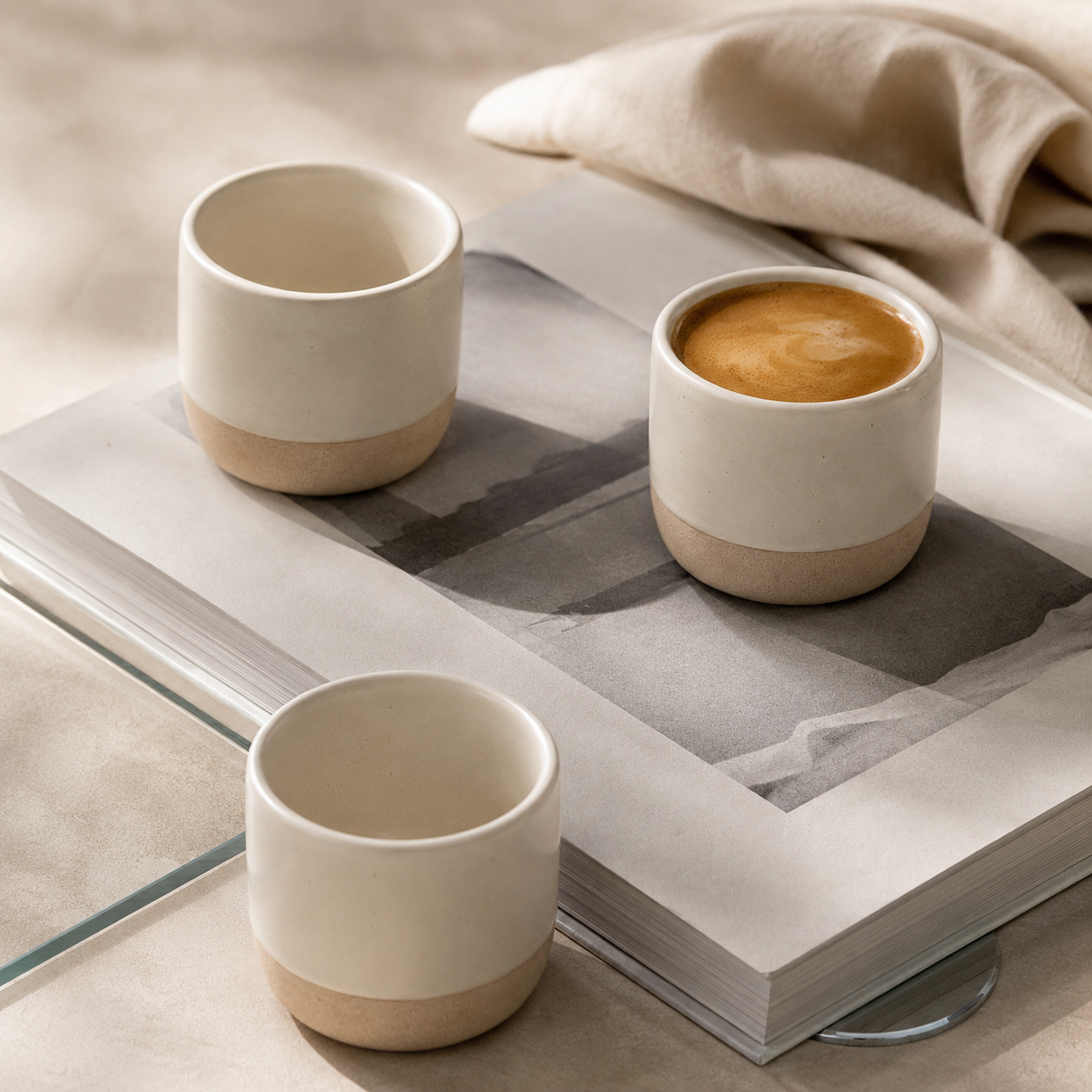 Three ceramic espresso cups with a two-tone cream and natural clay base, styled on an open book with a cup of coffee in one of the cups and a beige cloth in the background.