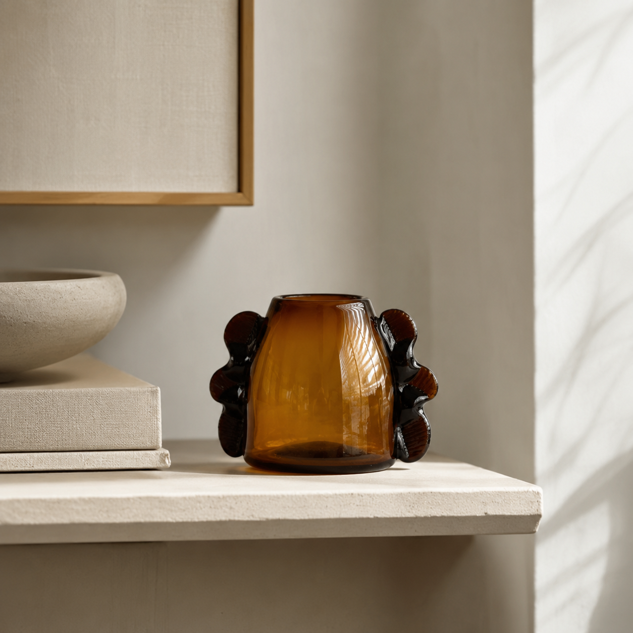 An amber glass vase with dark brown sculptural side details sits on a light beige shelf in a minimalist interior setting.