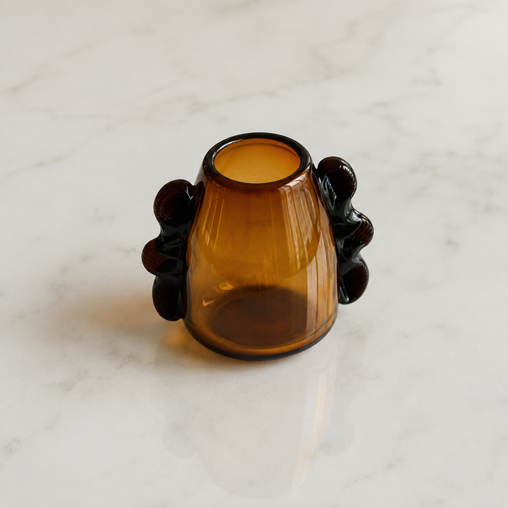 An amber glass candle holder with sculptural dark brown side handles, photographed on a white marble surface.