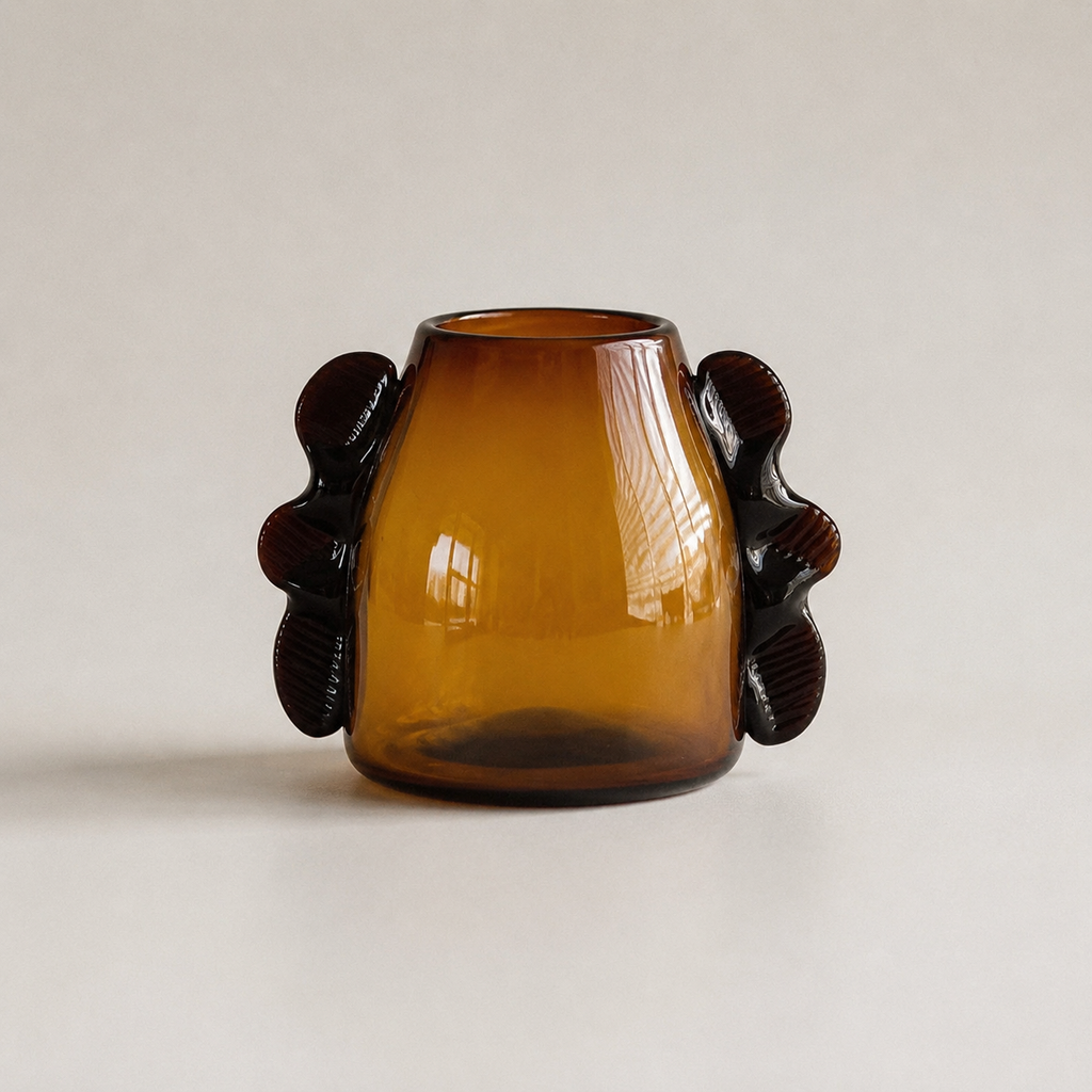 An amber glass vase with a rounded body and two dark sculptural handles on the sides, photographed against a plain light beige background.