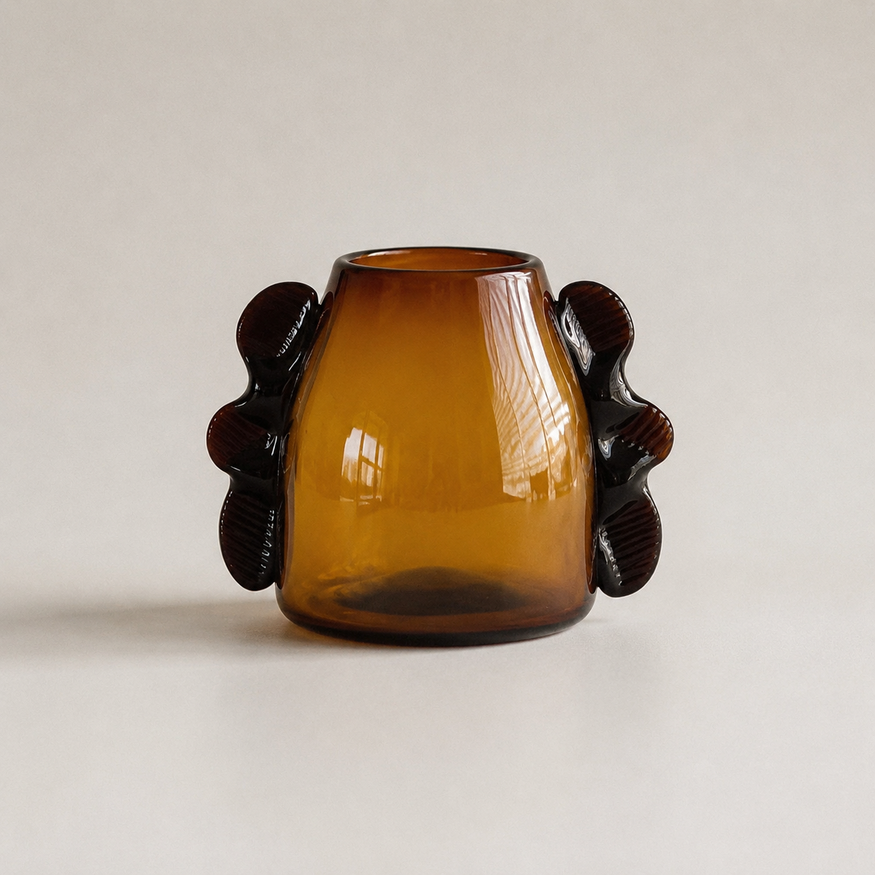 An amber glass vase with a rounded body and two dark sculptural handles on the sides, photographed against a plain light beige background.