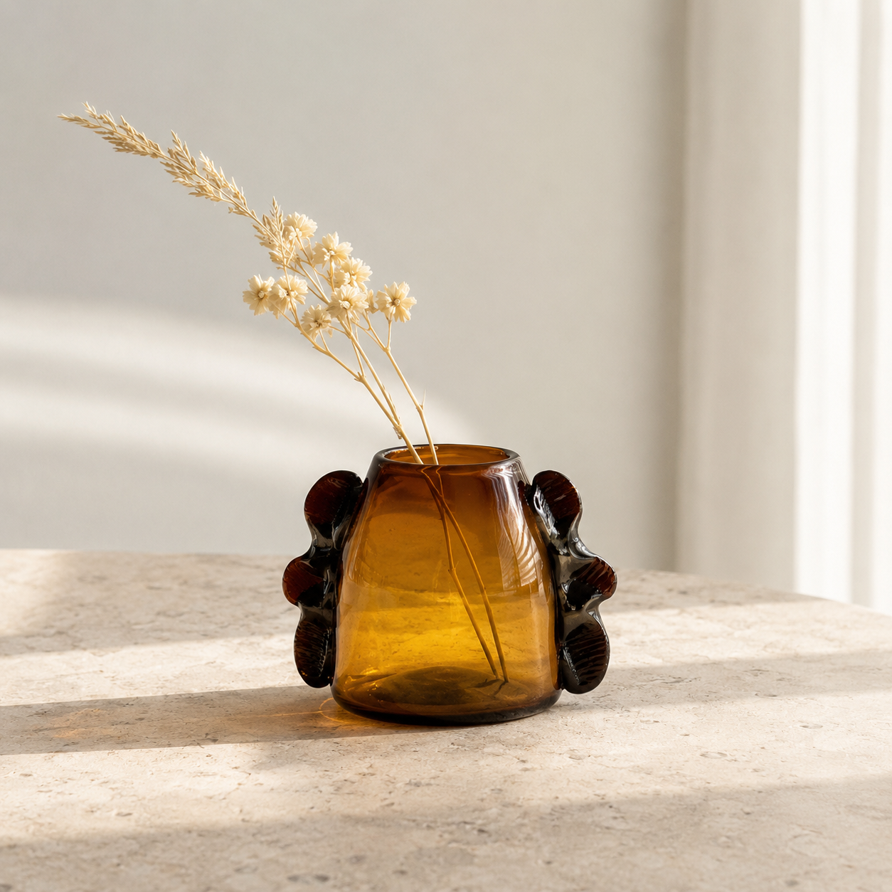 An amber glass vase with dark brown sculptural side details, holding dried beige flowers, set on a light stone surface against a soft neutral wall.