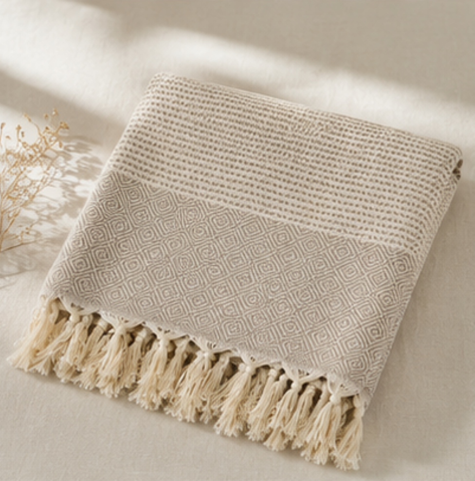A folded woven throw blanket with fringe tassels, shown on a light neutral fabric surface with soft natural shadows. The blanket has a geometric diamond pattern in warm beige and cream tones.