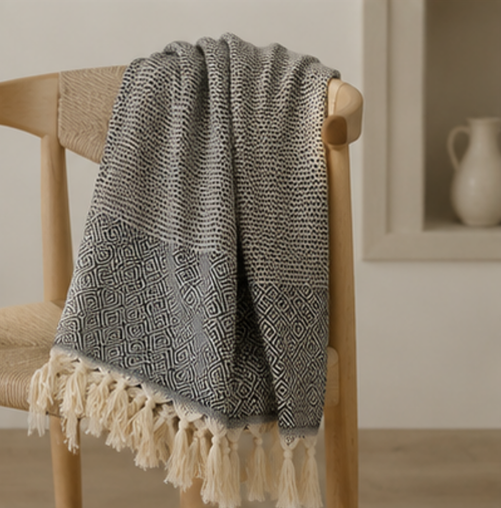 A gray and white patterned throw blanket with cream tassels draped over a light wood chair in a softly lit interior setting.