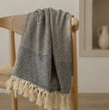 A gray and white patterned throw blanket with cream tassels draped over a light wood chair in a softly lit interior setting.