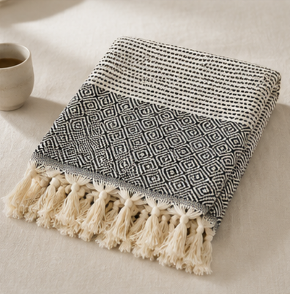 A folded woven throw blanket with cream tassels, shown on a light beige fabric surface. The blanket has a striped black-and-cream upper section and a geometric diamond pattern in black and cream across the middle, with a solid cream tassel fringe along one edge.