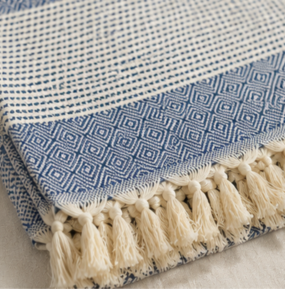 A folded woven textile with cream tassels, shown in close-up. The fabric features a cream and blue geometric diamond pattern and striped sections, with thick cream tassels along one edge.