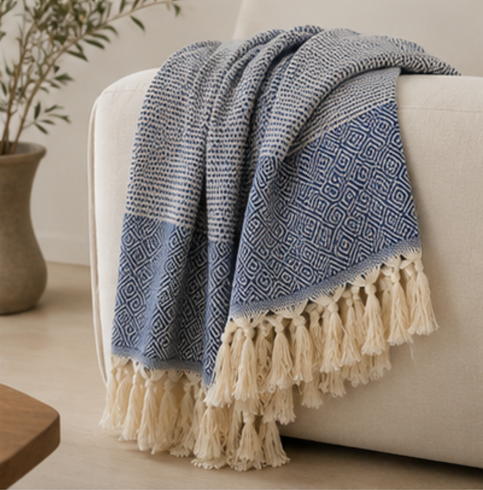 A blue and cream patterned throw blanket with tassels draped over a light beige armchair in a softly lit room.