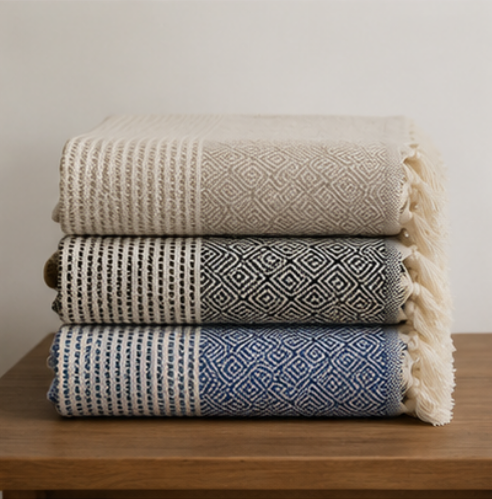 Three folded woven throw blankets stacked on a wooden surface against a plain light wall. The top blanket is cream with a geometric pattern and fringe, the middle is dark gray and white, and the bottom is blue and white.