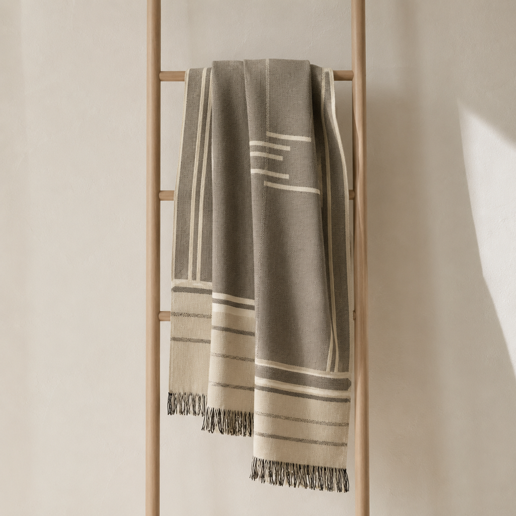 A woven throw blanket in taupe and cream with geometric stripe patterns and fringed edges, draped over a minimalist wooden ladder against a warm beige wall.