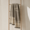 A woven throw blanket in taupe and cream with geometric stripe patterns and fringed edges, draped over a minimalist wooden ladder against a warm beige wall.