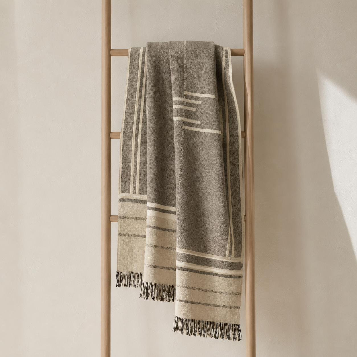 A woven throw blanket in taupe and cream with geometric stripe patterns and fringed edges, draped over a minimalist wooden ladder against a warm beige wall.