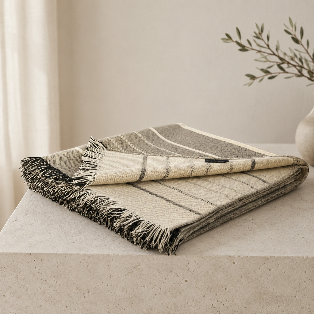 A folded woven throw blanket with fringe, shown on a light stone surface in a softly styled interior. The blanket has a striped pattern in warm beige, cream, taupe, and gray tones.